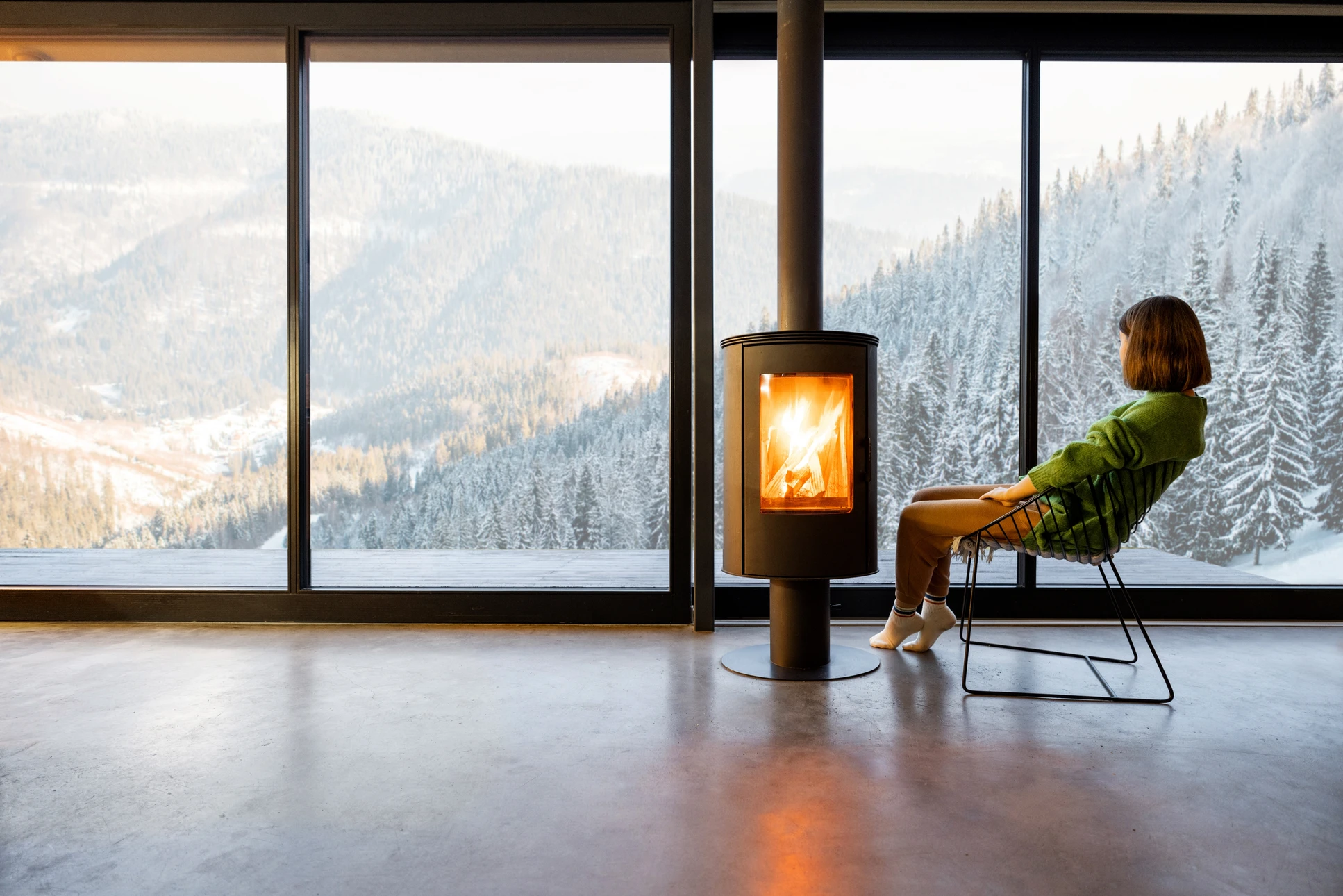 Femme assise sur chaise design face à poêle à bois moderne dans salon contemporain avec baies vitrées panoramiques donnant sur paysage montagneux enneigé