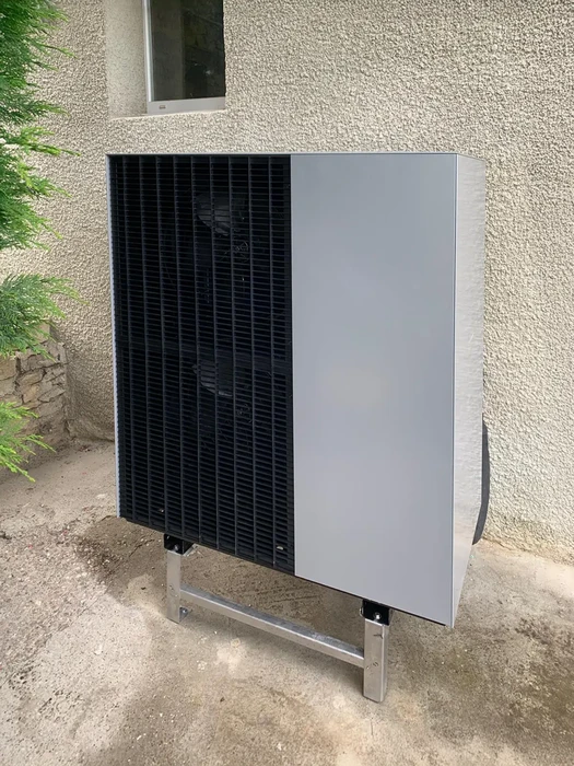 Extérieur résidentiel avec unité de climatisation installée contre mur crépi beige, végétation visible en arrière-plan
